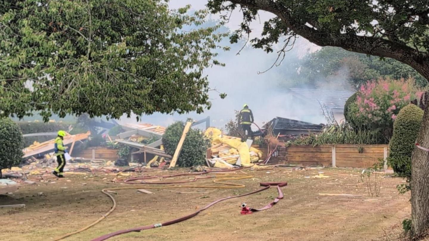 A picture from far away from the scene. There is the house spread out everywhere you can see wood and metal lying all over the floor. To the right of the image, you can see a large tree.