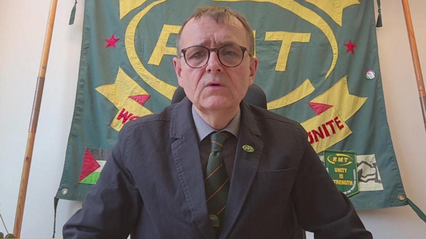 The RMT's John Leach wearing a blue jacket and red and green striped tie, sitting in front of a RMT banner.