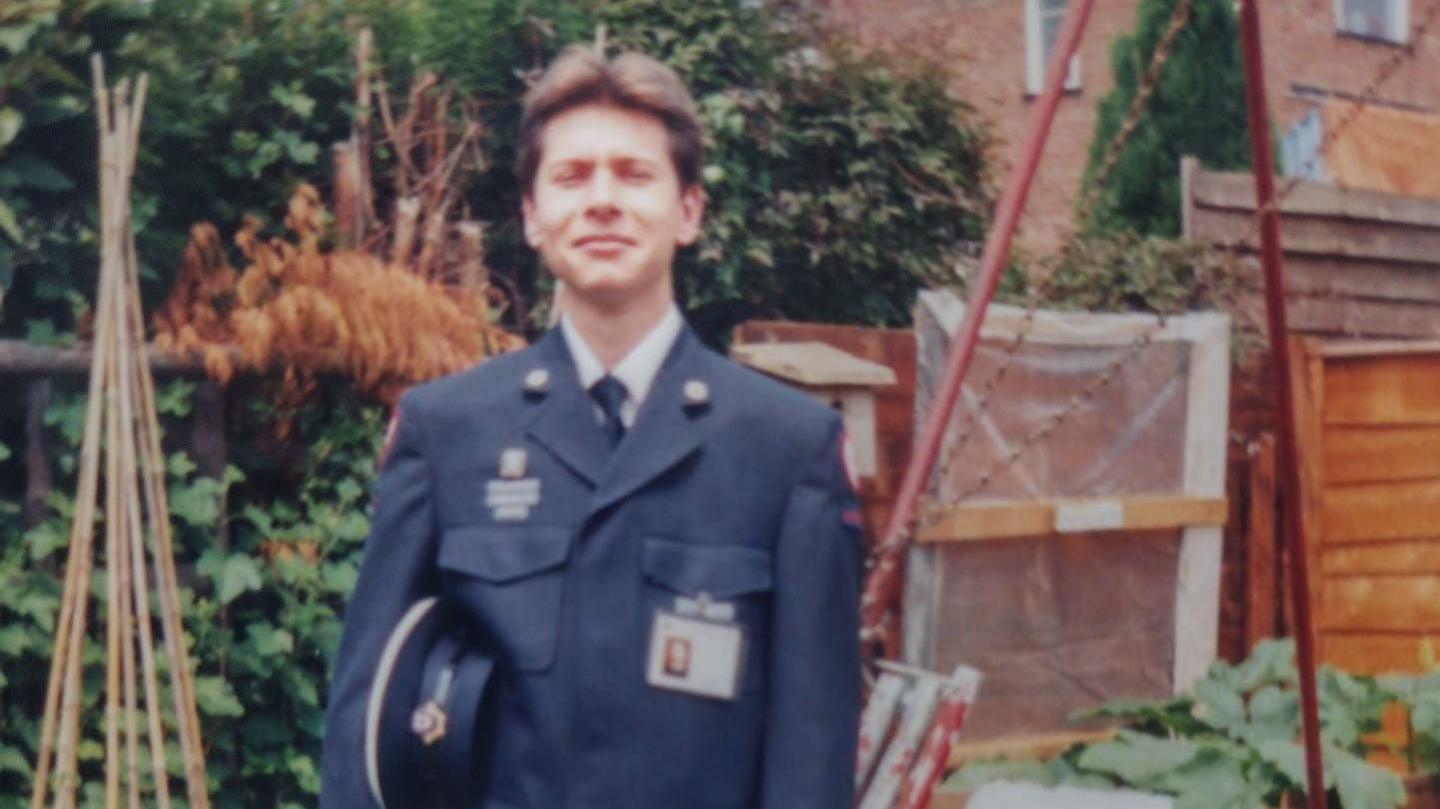 A young man is standing in a garden with a blue uniform on. He has a blue tie and blue jacket with some sort of identity badge on the chest. He has a cadet hat under his right arm.