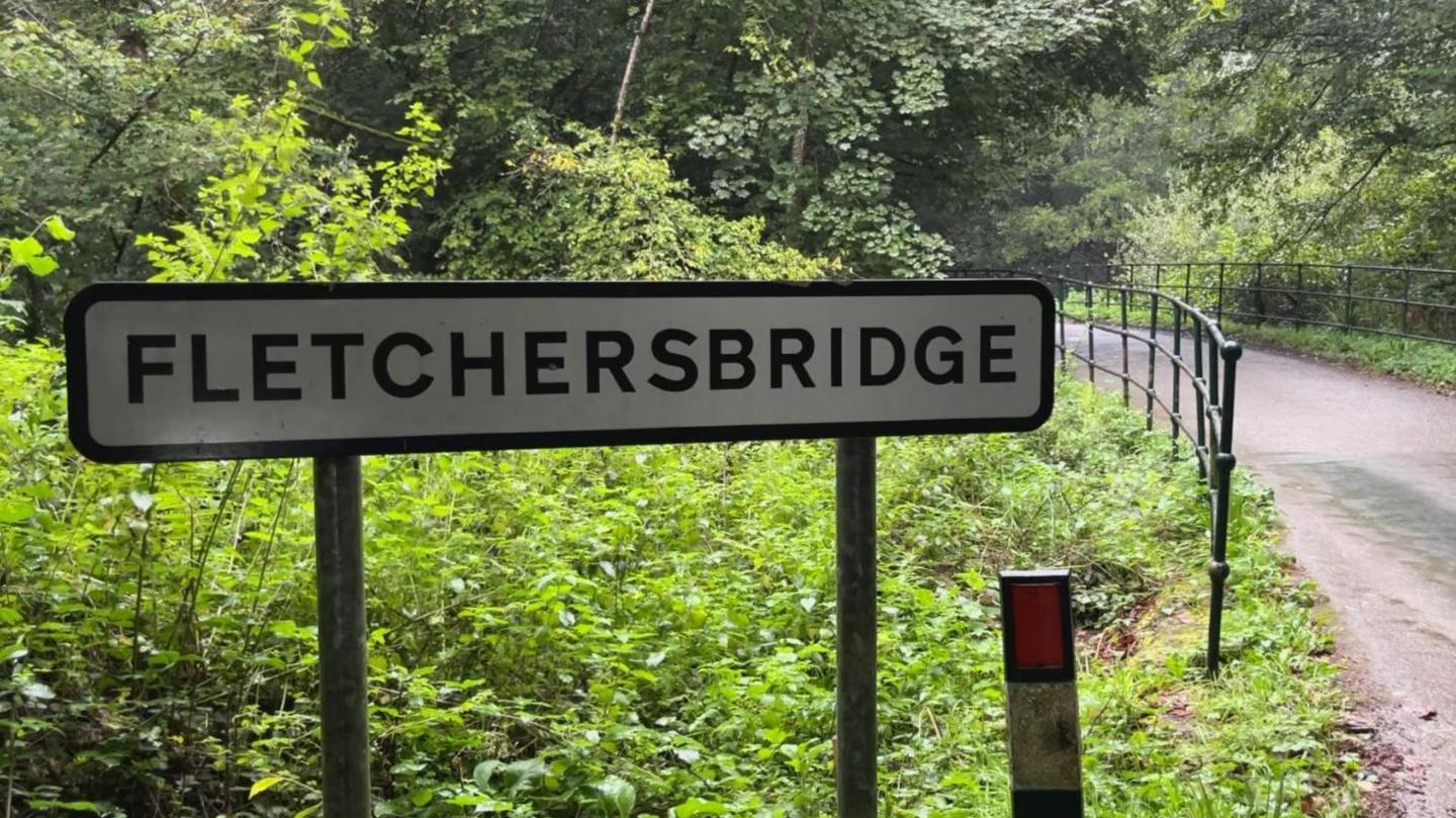 A sign for Fletchersbridge where the incident took place. The sign is next to a road with a railing and bridge. Vegetation and trees surround the sign.