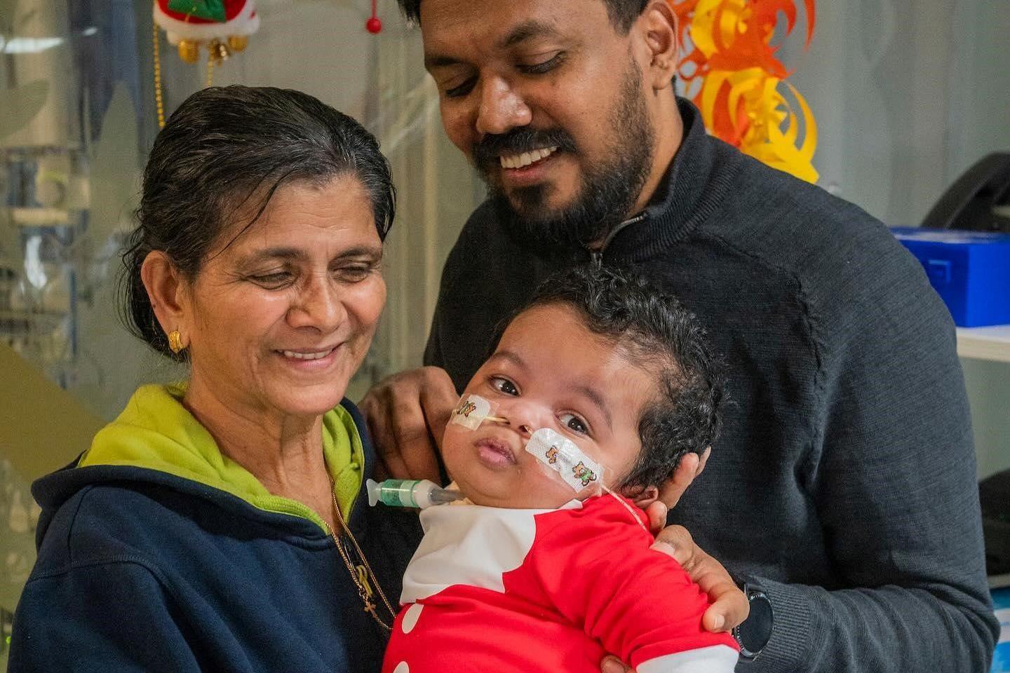 Gabriel's granny is wearing a blue hooded top and has black hair tied up and gold earrings. She is looking at her grandson while holing him in her arms and smiling. Gabriel is wearing a red Santa outfit and has tubes in both nostrils that are Sellotaped to his face. His dad is in the background in a dark top and smiling while looking at his son.