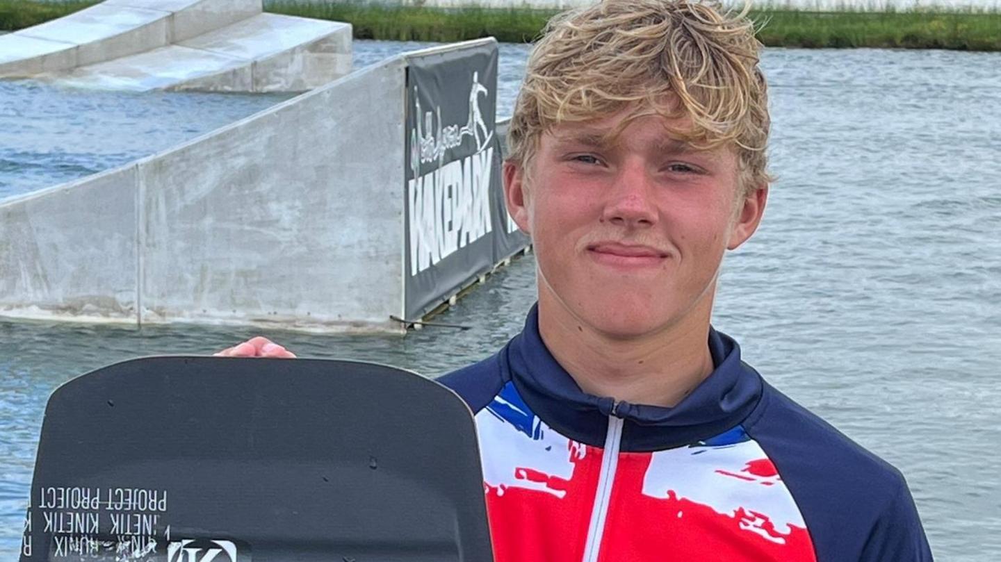 This picture shows a person standing near water, holding a wakeboard. The individual is wearing a jacket featuring the Union Jack. In the background, there are ramps and structures typical of a wakeboarding cable park.