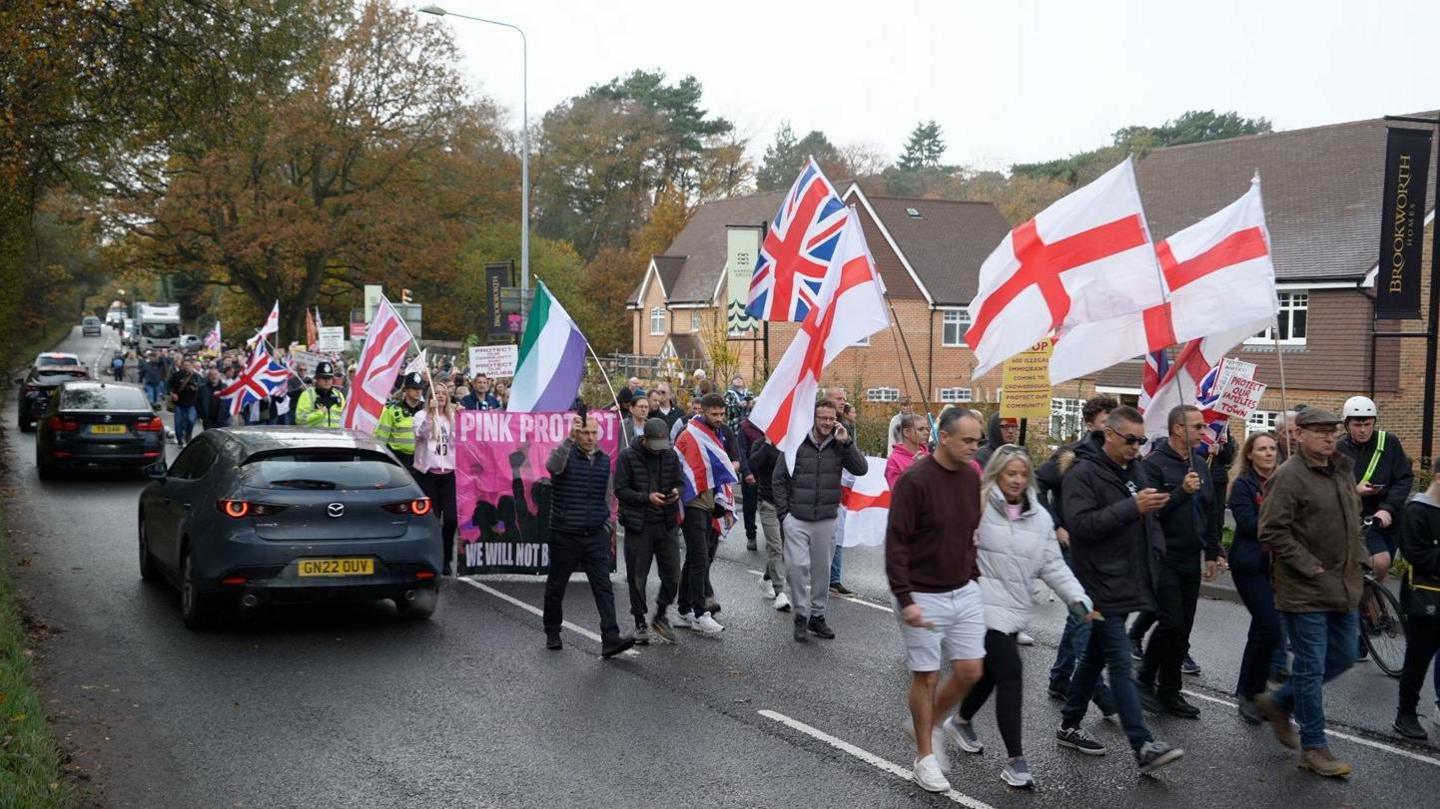 Hundreds of people holding St George flags and Union Jack flags marching along a road. There are cars driving by.