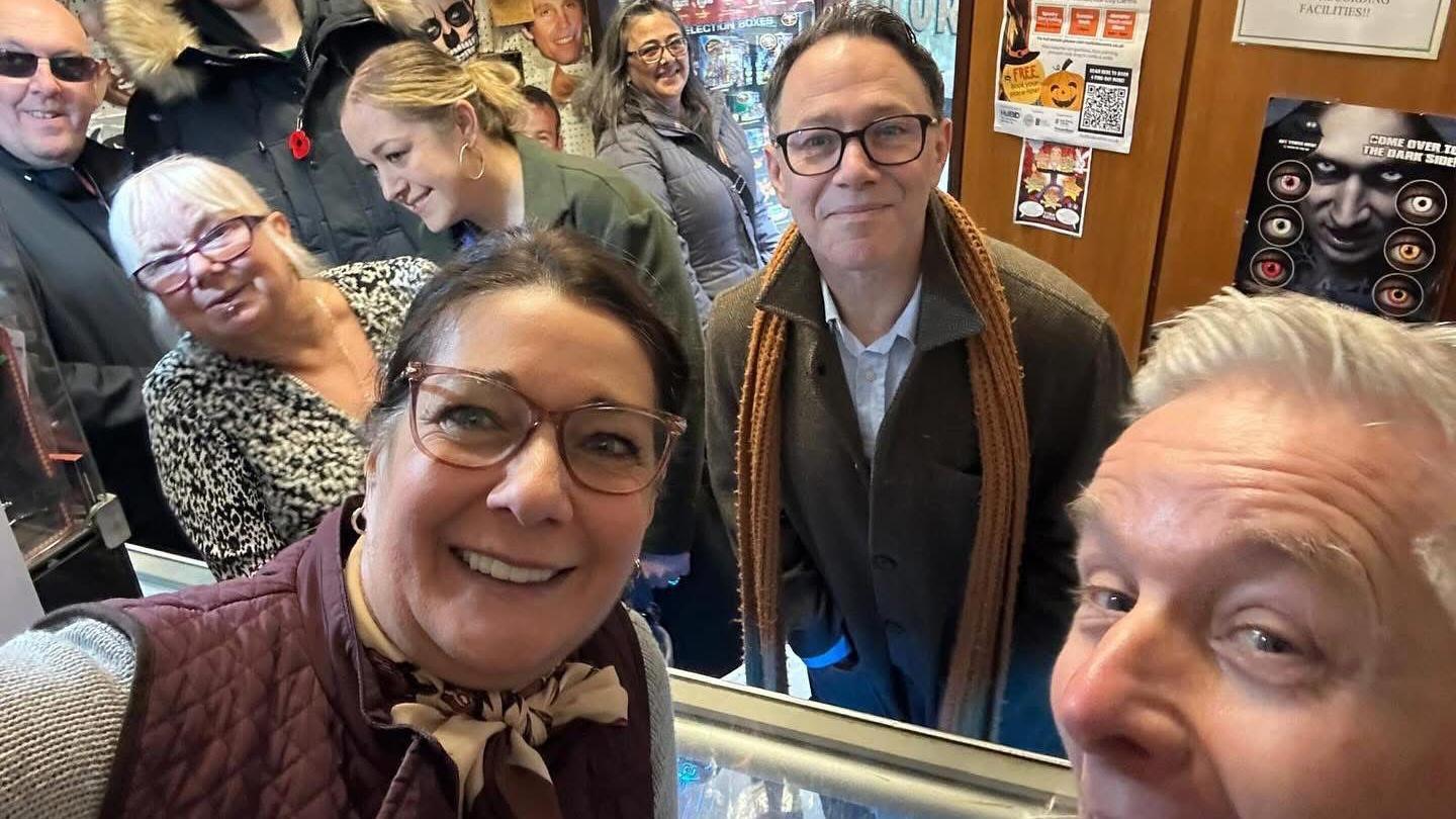 A group selfie with a mixture of males and females spread throughout in a small joke shop. A brunette female with large framed glasses and a burgundy gillete takes the photo and smiles. Behind her there are two males and three females who are leaning to and away from the camera, their heads are partially or fully cut out of the frame. To the right is a dark-haired man with black glasses, a khaki coat and mustard coloured scarf - he leans towards the camera. A white-haired man is partially cut out of frame, he has his mouth open wide.
