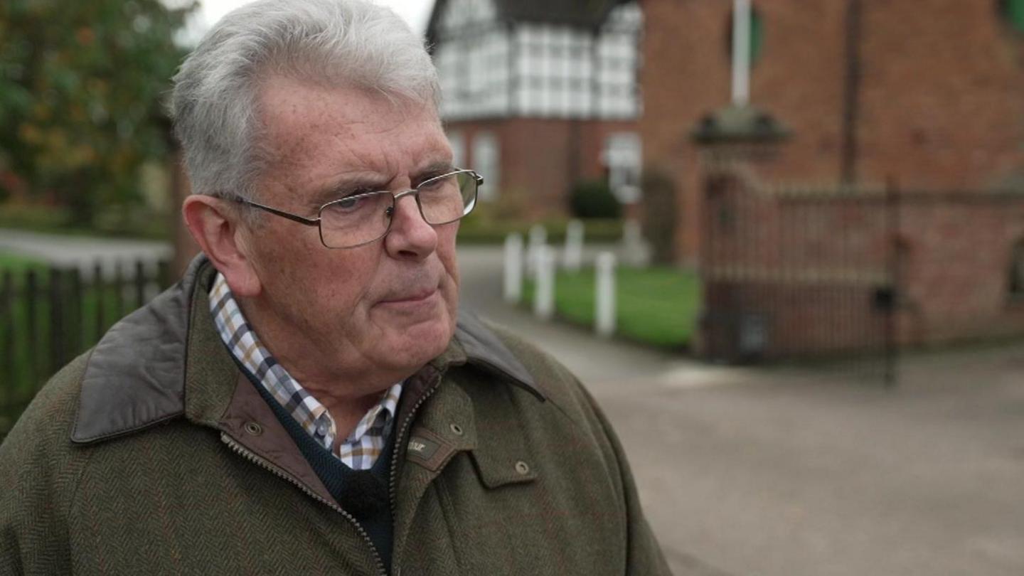 John Edge with grey wavy hair, glasses and a khaki overcoat. His unsold farmhouse is in the background.