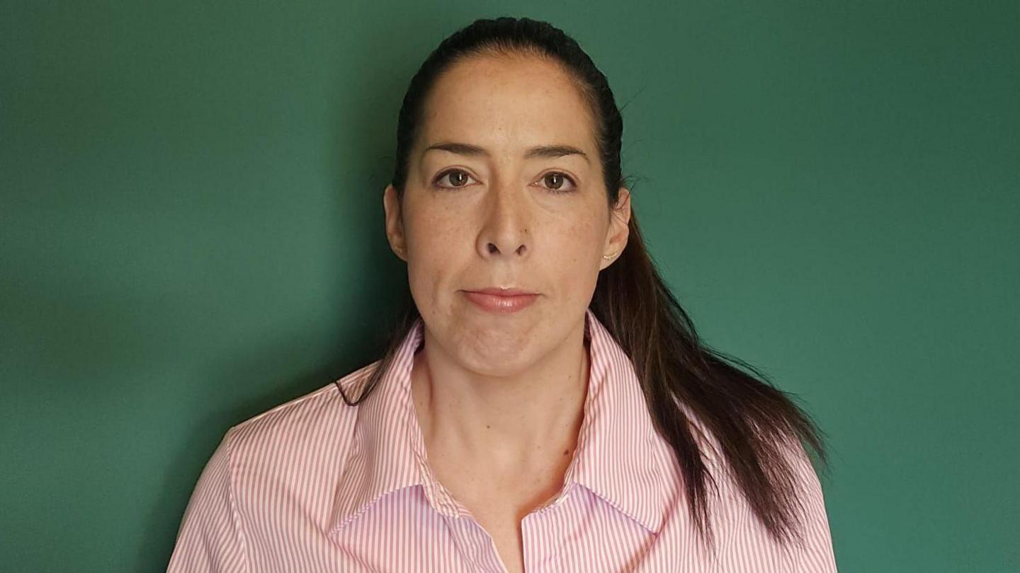 A woman with brown hair looks at the camera. She is wearing a pink and white shirt, and is standing in front of a dark green wall. 