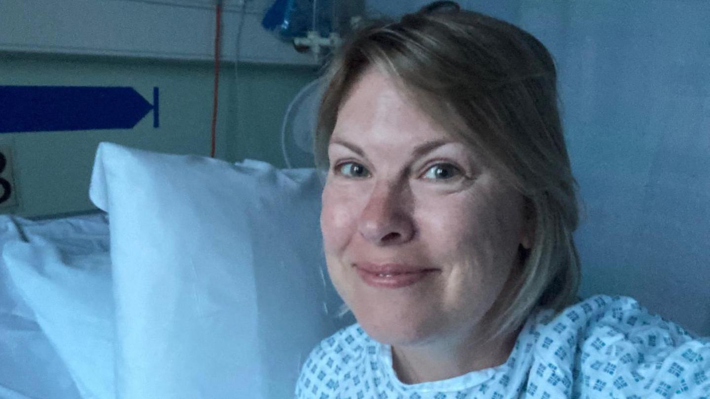 Woman smiles in a hospital gown on a bed in a clinical room with white pillows, curtain and medical equipment.