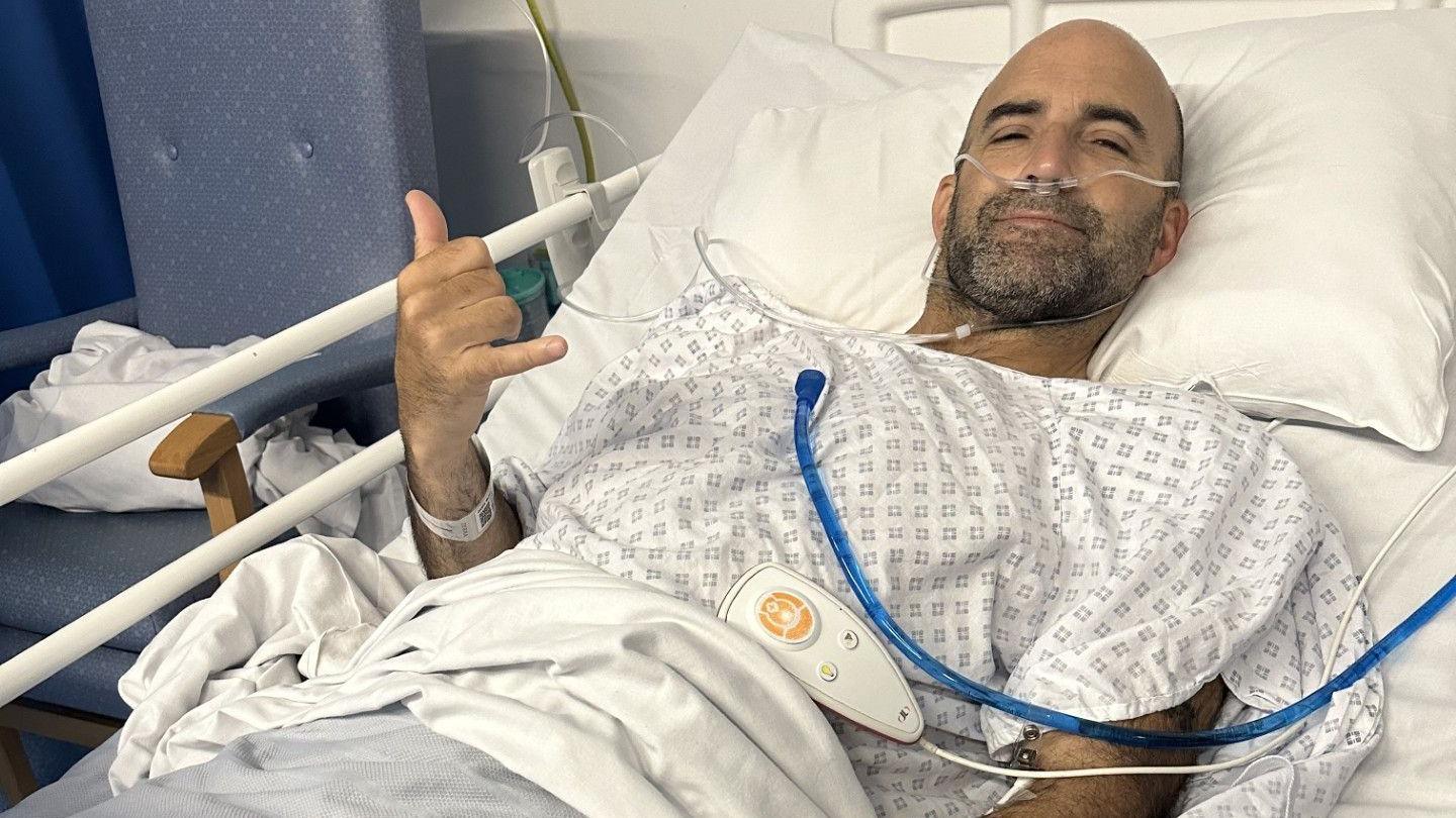 Michael Oakes laying in a hospital bed with tubes in his nose. He is smiling at the camera and making shaka gesture with his right hand