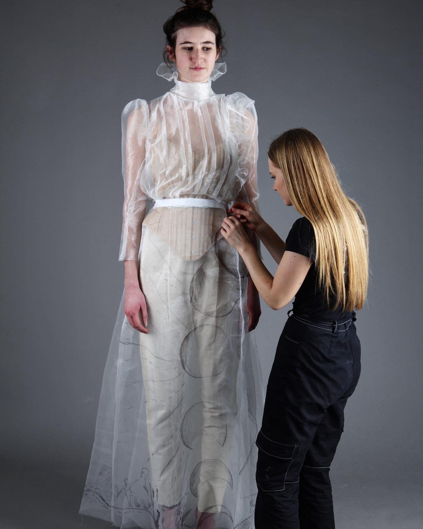 Evie standing in front of female model wearing period white lace floor length dress, making adujustments to the costume