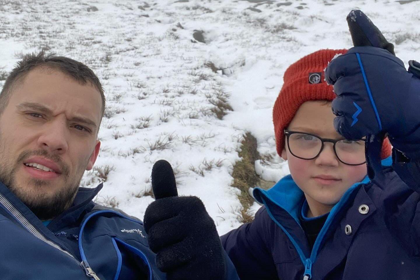Jake on the left and Rio on the right in the snow. Both are wearing thick blue coats and Rio a red woolly hat. They are holding their gloved thumbs up to the camera.