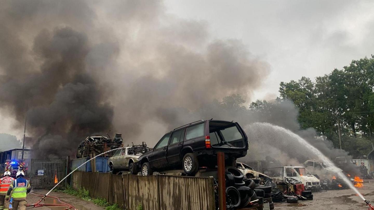 A car scrap yard with tyres and cars stacked on top of each other next to a wooden fence. Smoke is billowing into the sky and flames can be seen in two separate areas of the yard. In the foreground are two firefighters and two hoses spraying water towards the vehicles