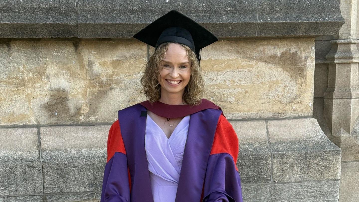 A woman in a graduation gown smiles at the camera. She is wearing a black mortarboard and a purple and red graduation robe. Beneath that she is wearing a lilac outfit. She has curly dark blonde hair. Behind her is a stone wall.