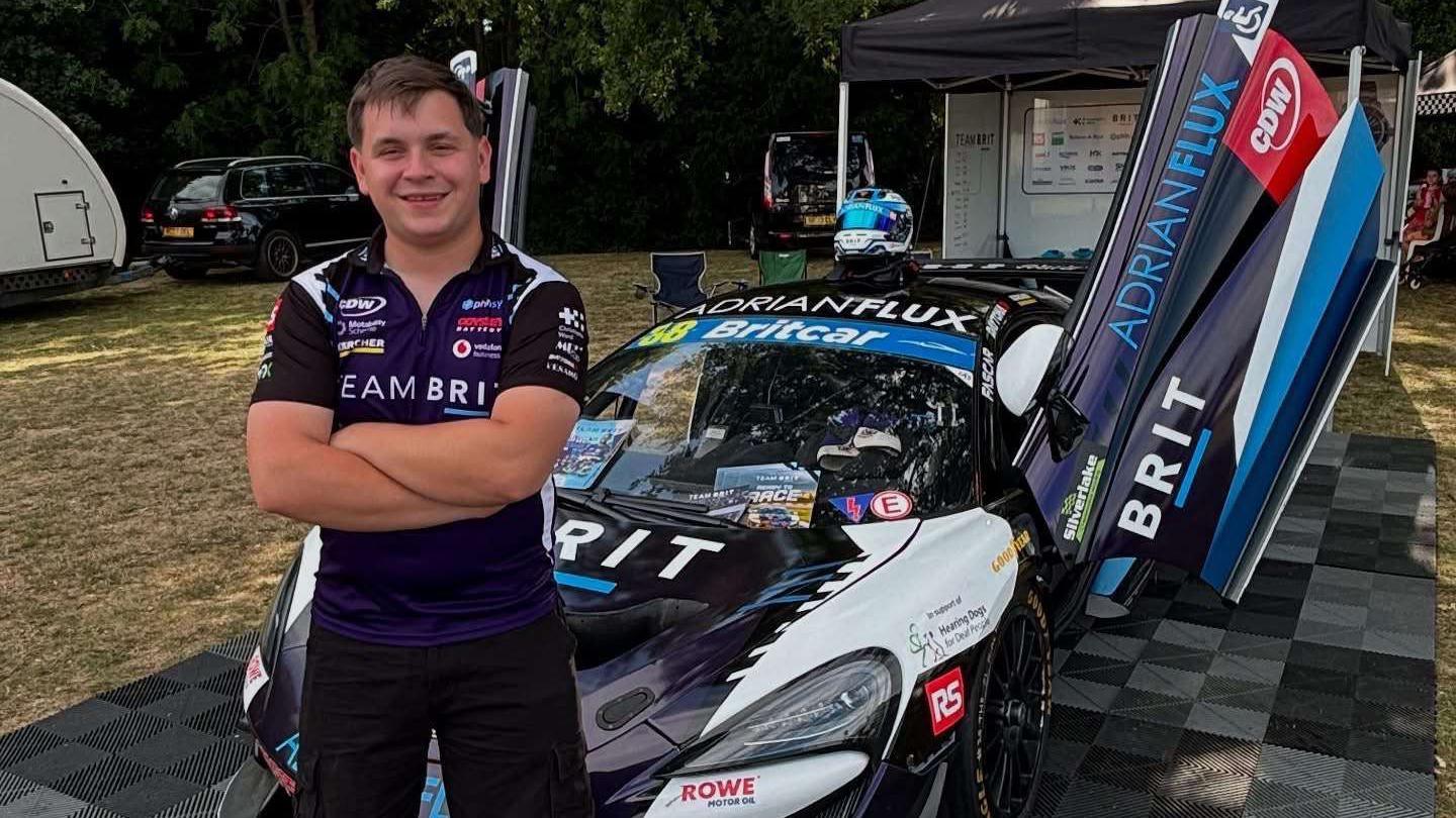 A smiling man in a Team BRIT shirt and shorts stands in front of a McLaren race car with its doors open, parked on a checked mat at a race paddock surrounded by trees.