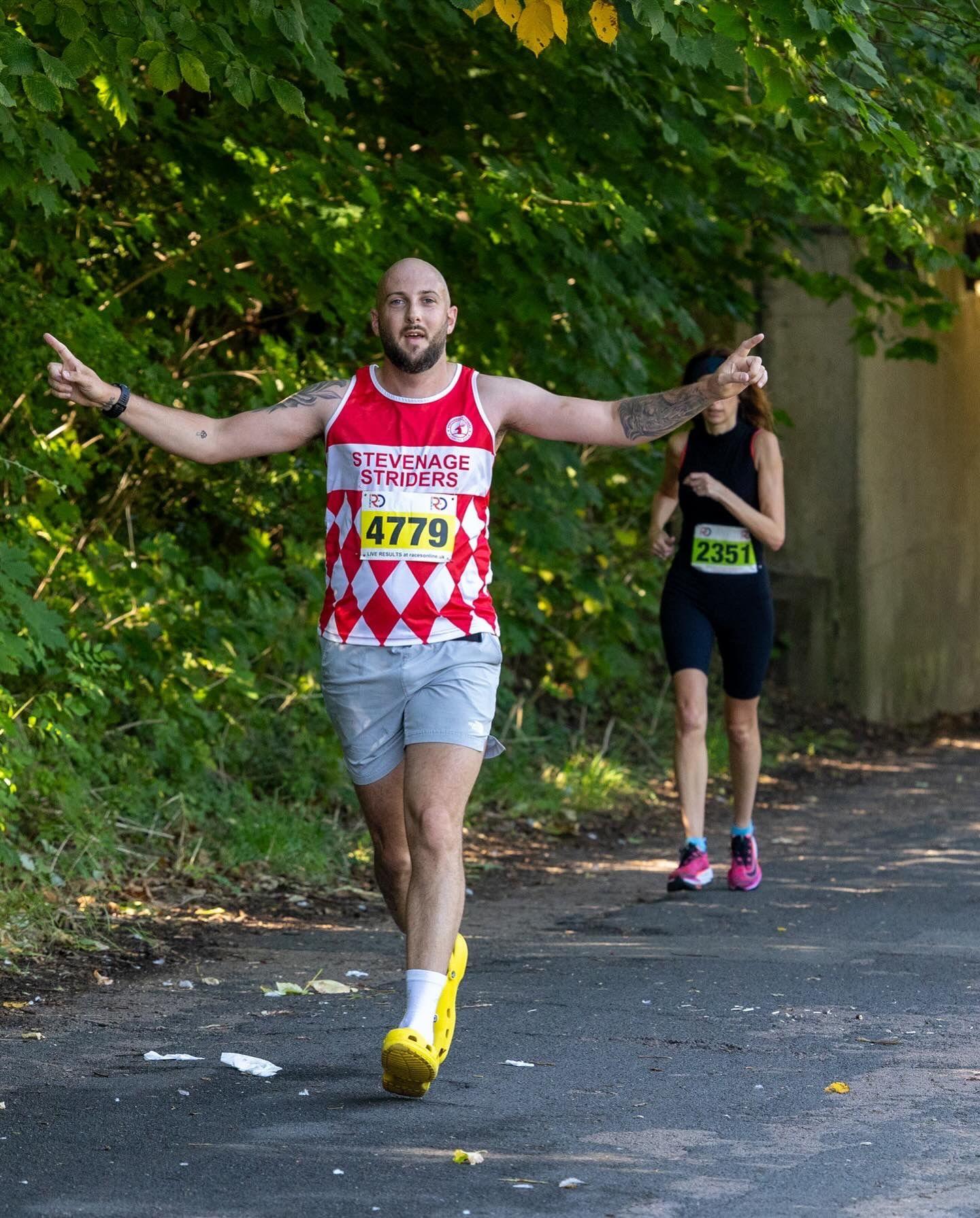 Daniel, holding two arms out, running, in yellow plastic shoes, wearing grey shorts, and a red and white running top. You can see a grass verge and a runner behind him.