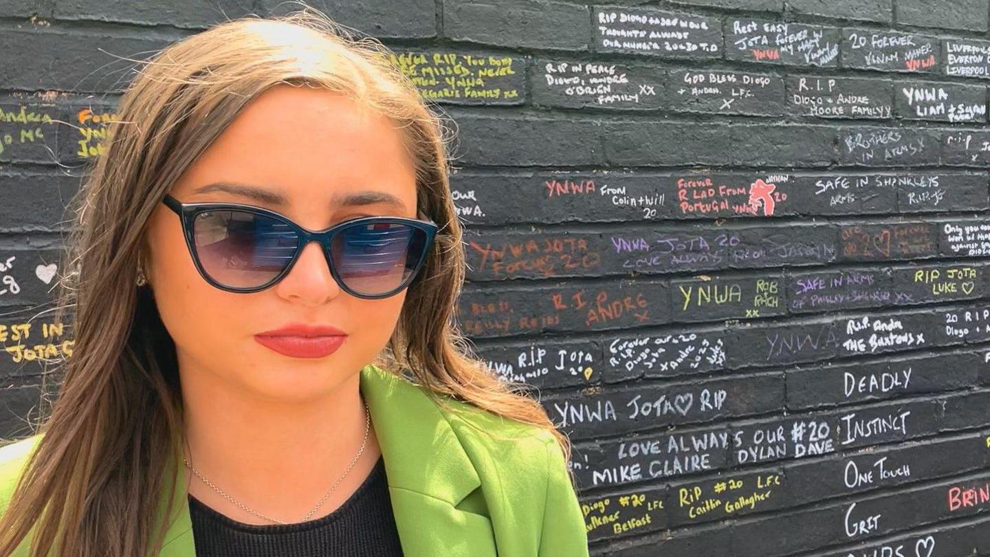 Grace Merritt, 22, who has long straight light brown hair and is wearing sunglasses, a green blazer and a black top, stands in front of a black wall covered in messages written in different colours.