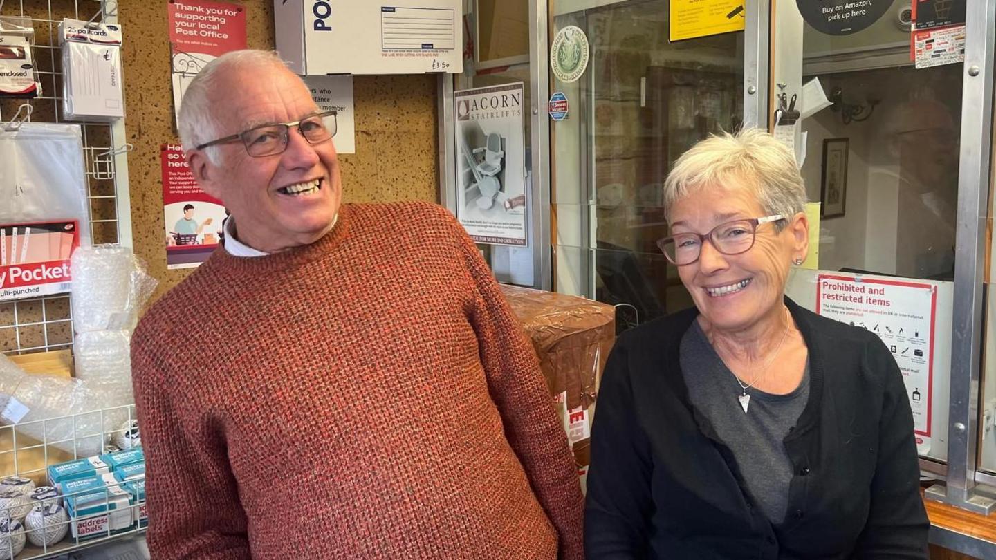 Phil Dolby, pictured with white hair, an orange jumper and glasses, next to ex-wife Karen Woodward, who has white hair, a black cardigan and glasses inside Crich Post Office