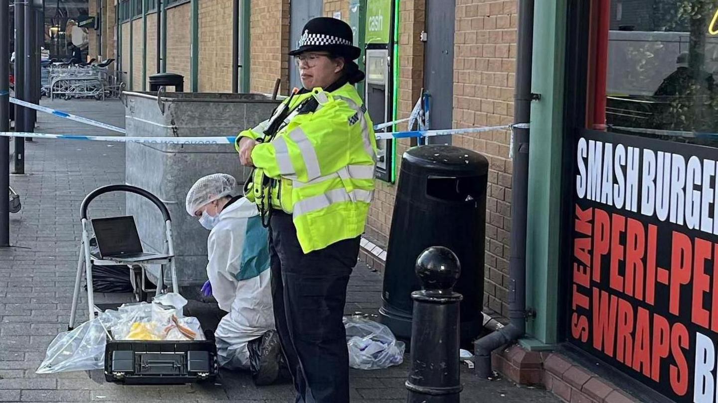 A woman police officer wearing a yellow coat stands with folded arms outside a fast food shop. Kneeling to her right is a crime scene investigator dressed in a white suit.


