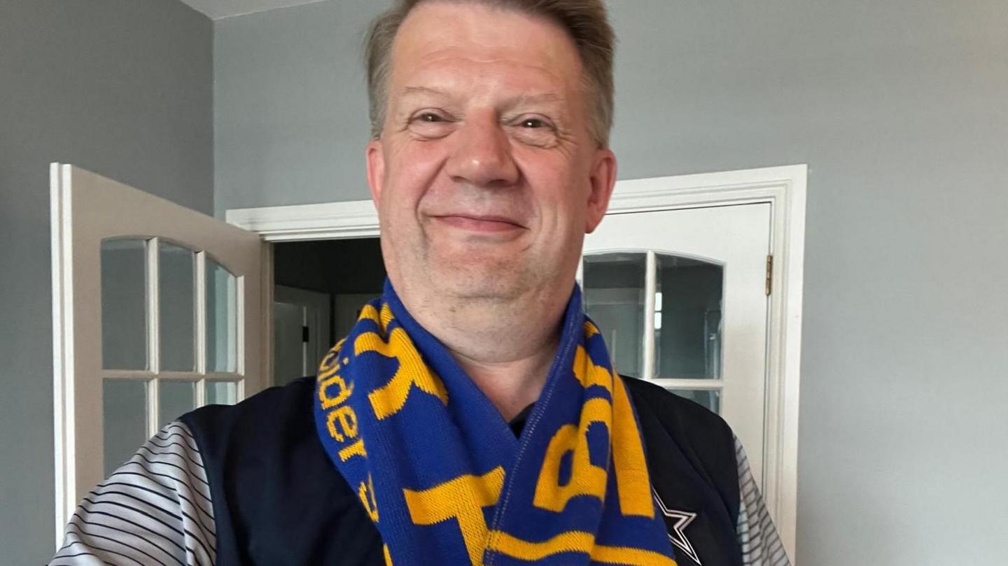 Cameron McKay has short brown hair and is seen smiling.
He is standing in a hallway with a white doorframe behind him. 
He is wearing a striped navy and white top, as well as a yellow and blue Bangor scarf.