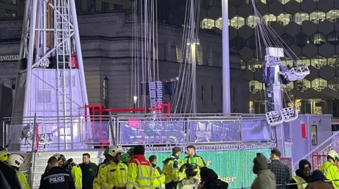 A broken fairground ride with part of it dangling by its cables and lots of people in yellow jackets standing around the bottom