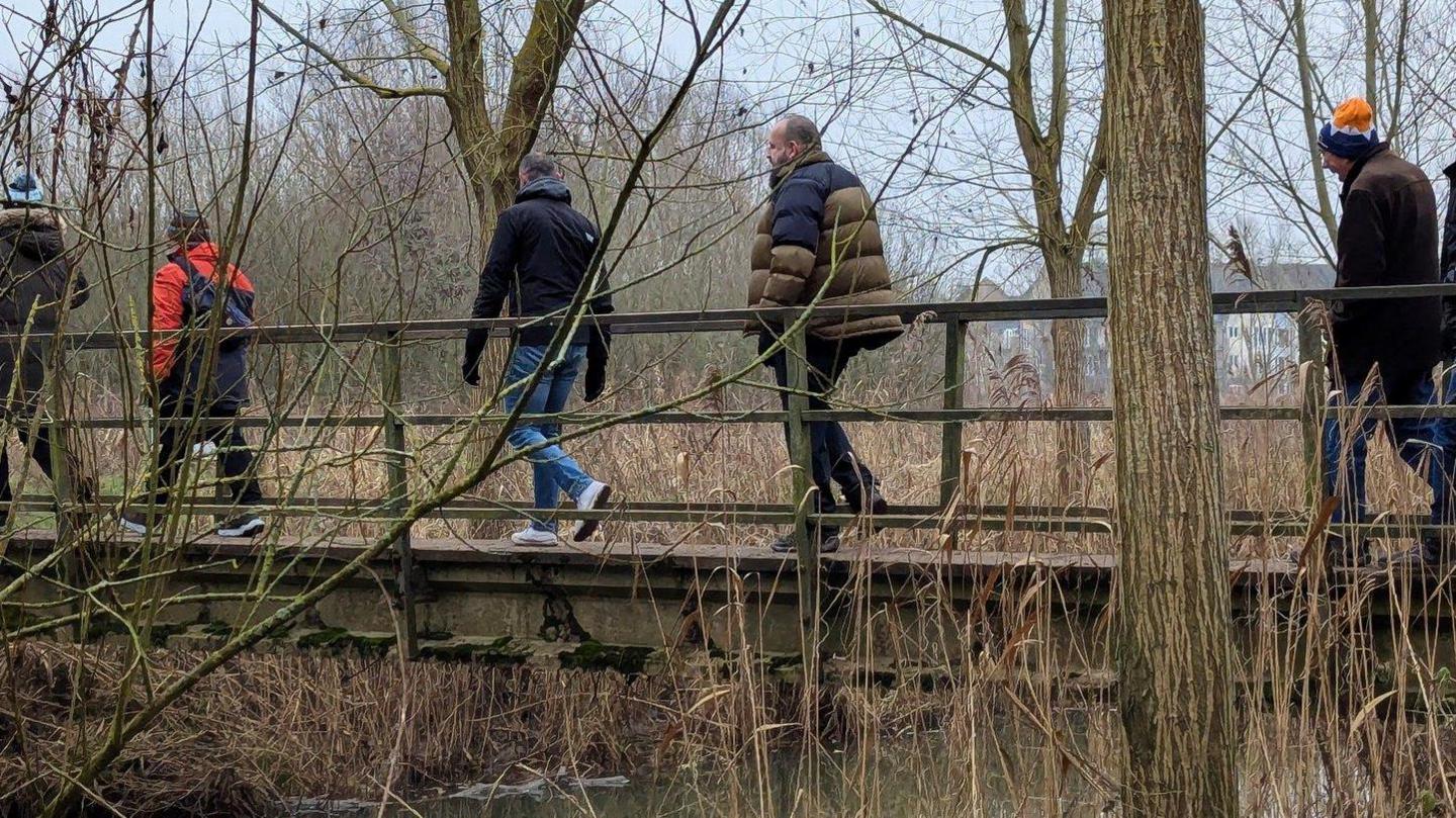 Six men are walking in pairs across a footbridge in a woodland area. They are taking part in Walk & Talk 4 Men St Neots. They wear coats, some have hats and the weather looks cold and grey. There are no leaves on the surrounding trees.   