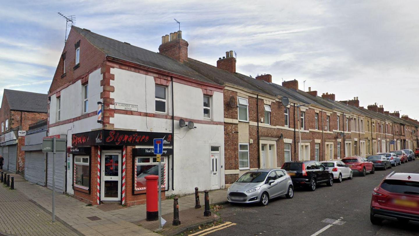 Claremont South Avenue, in Bensham, Gateshead. Cars are parked outside a row of terraced houses. At the end of the street is a barbers. A red post box stands outside the barbers.