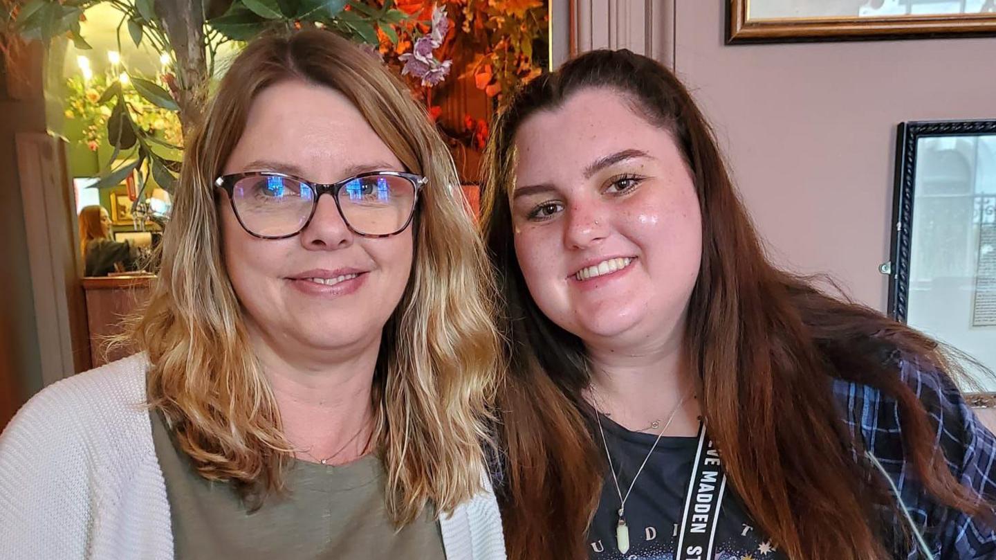 Shelley Macpherson and Beth Langton smiling and looking into the camera. Shelley is wearing an olive green top, a cream cardigan and glasses. Beth is wearing a dark T-shirt with a blue checked shirt over the top