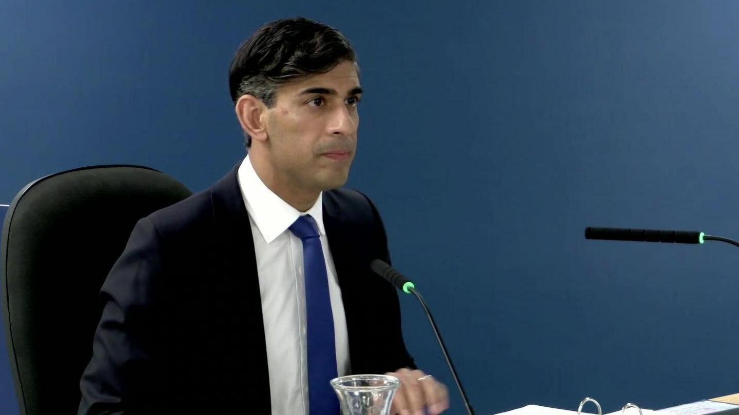 Rishi Sunak sitting down at the Covid inquiry giving evidence. He is wearing a dark-blue suit with a white shirt and royal blue tie. 