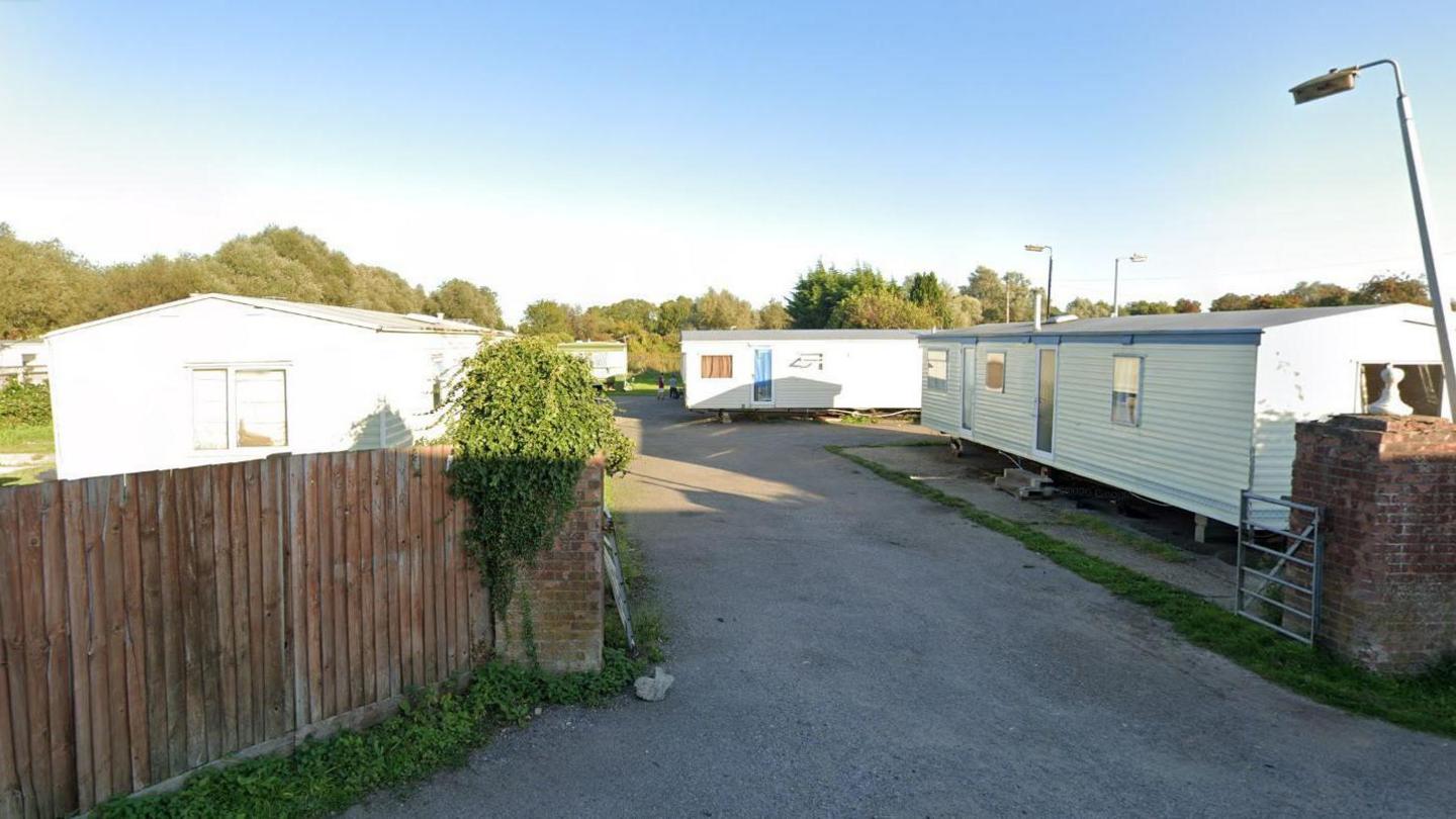 A road leading into a caravan park. There is a wooden fence on the left and brick pillars by the gate. Four mobile homes are seen in the sunlight, with trees and street lamps behind them.