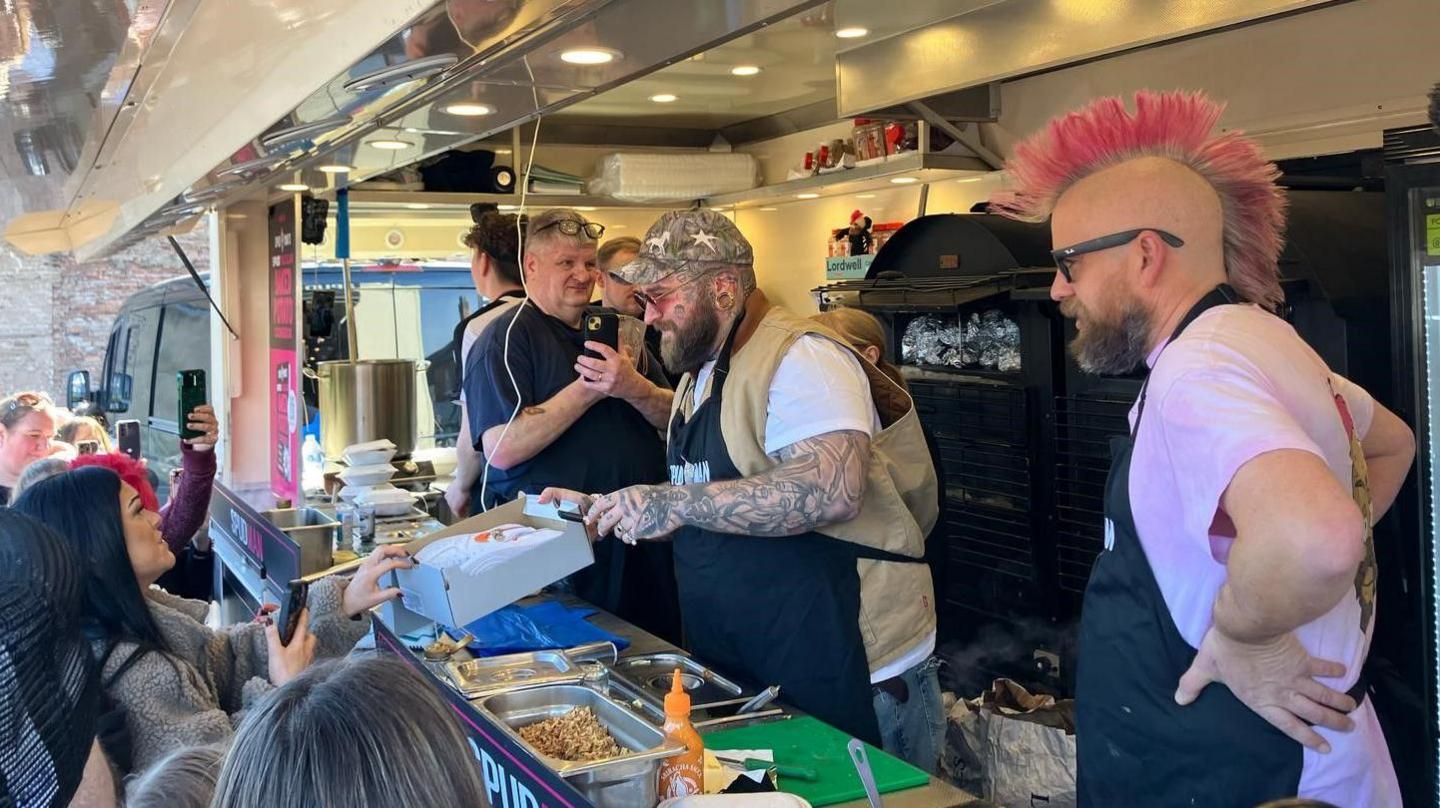 A heavily tattooed man passes a cardboard box with food containers in to lady with long, black hair. A man with a pink mohawk is standing next to him with his hands on his hips. They are both wearing black aprons as they are in a street vendor truck. 