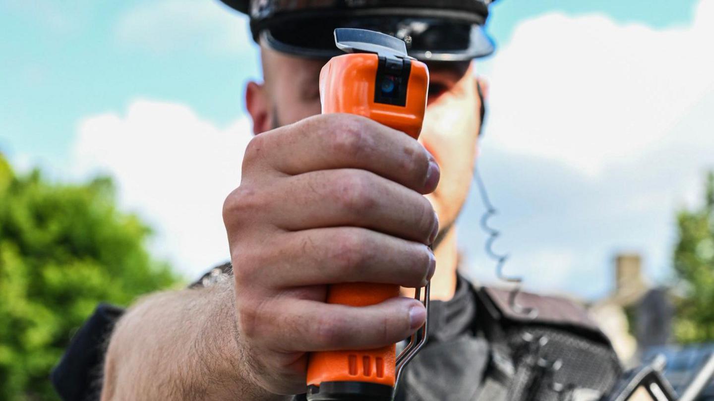 A police officer man holding an orange spray device in his hand - the device looks like a futuristic deodorant can.