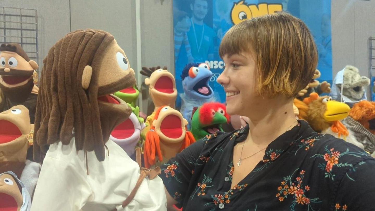 Hannah Fleming-Hill with short blond hair wearing a dark top with orange flowers, holds a puppet with brown hair and beard, wearing a white cloak.  There are other puppets with open mouths behind them