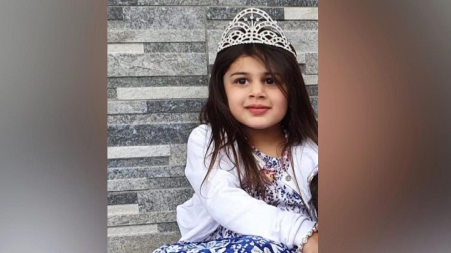 A little girl with big brown eyes and dark brown hair flowing over her shoulders is wearing a silver tiara, floral patterned blue and white dress and white cardigan. There is a gentle smile on her face.