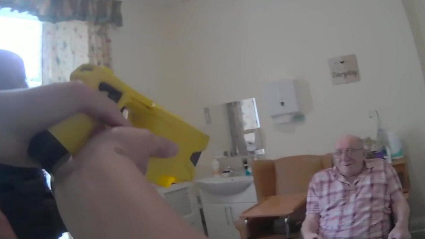 Donald Burgess sits in a chair beside his bed in a care home. He is wearing a red and white checked shirt. One police officer is visible to the left side of the picture and the hands of another, pointing a yellow taser at him, are also visible to the left side of the screen.