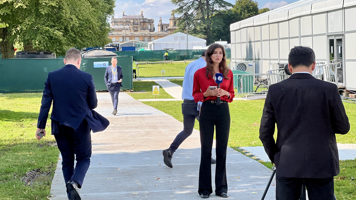 A woman presenter holding a microphone getting ready to go on air, with journalists milling around temporary structures with Blenheim Palace in the background 