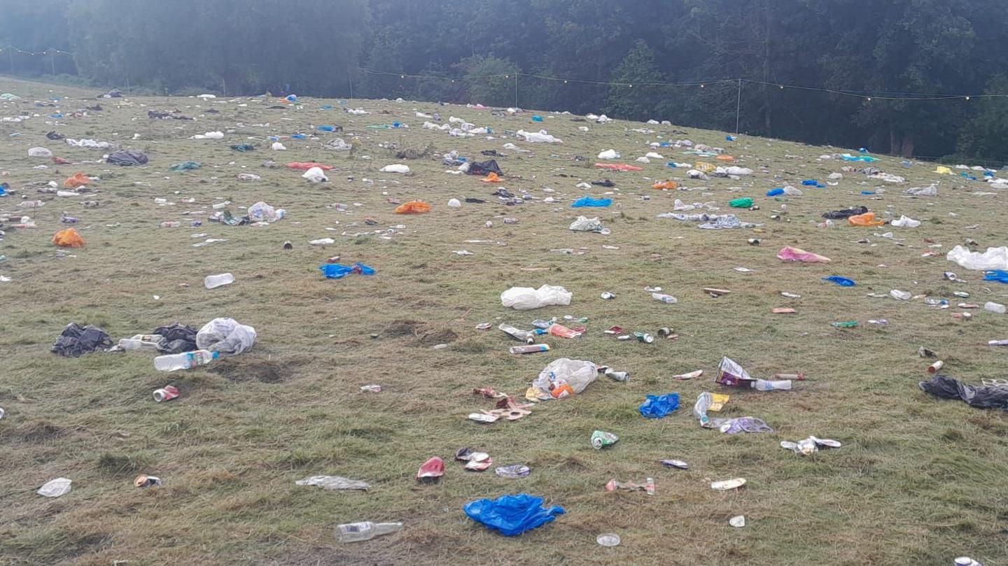 A grassy field strewn with litter including empty cans, plastic bottless, carrier bags and other debris.