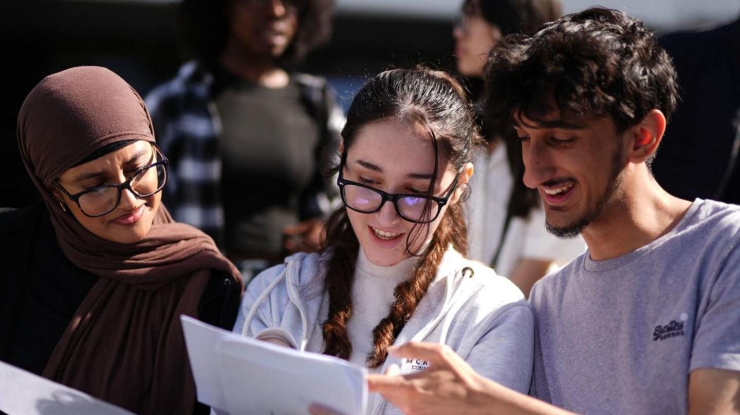 Two female students and a male student look down at an exam results sheet they've just opened. 