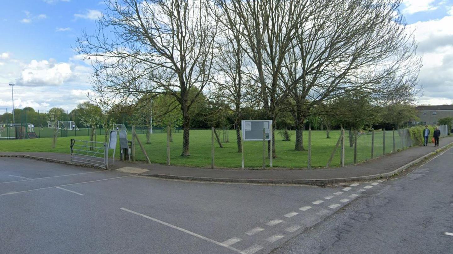 A Google maps screenshot of the driveway into a sports ground. Football goalposts are visible on the far left behind a metal fence and there is a grassy patch ahead with several trees dotted around next to the road.