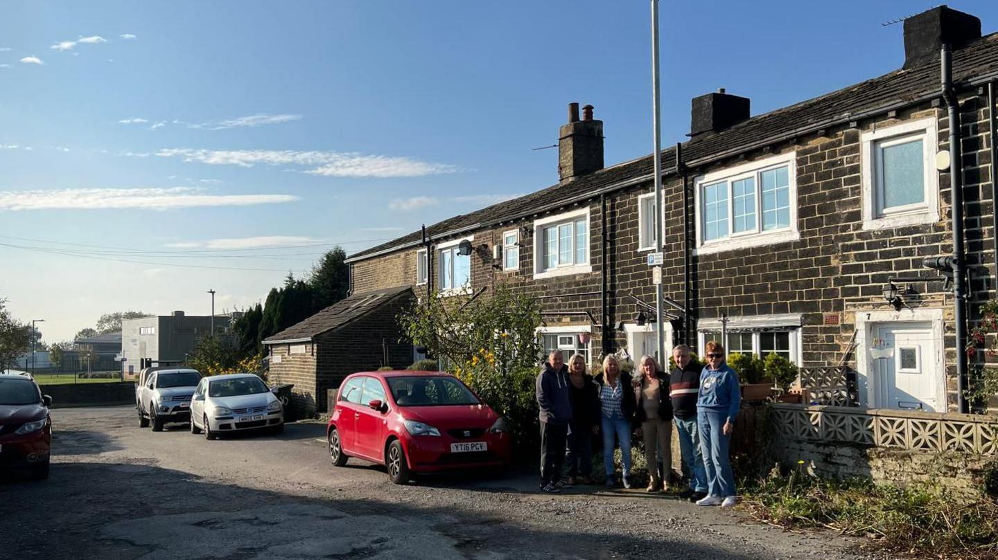 Six people standing under a 'residents only parking' sign with a big street scene surrounding them. There are several cars parked around them anda row of houses behind them.