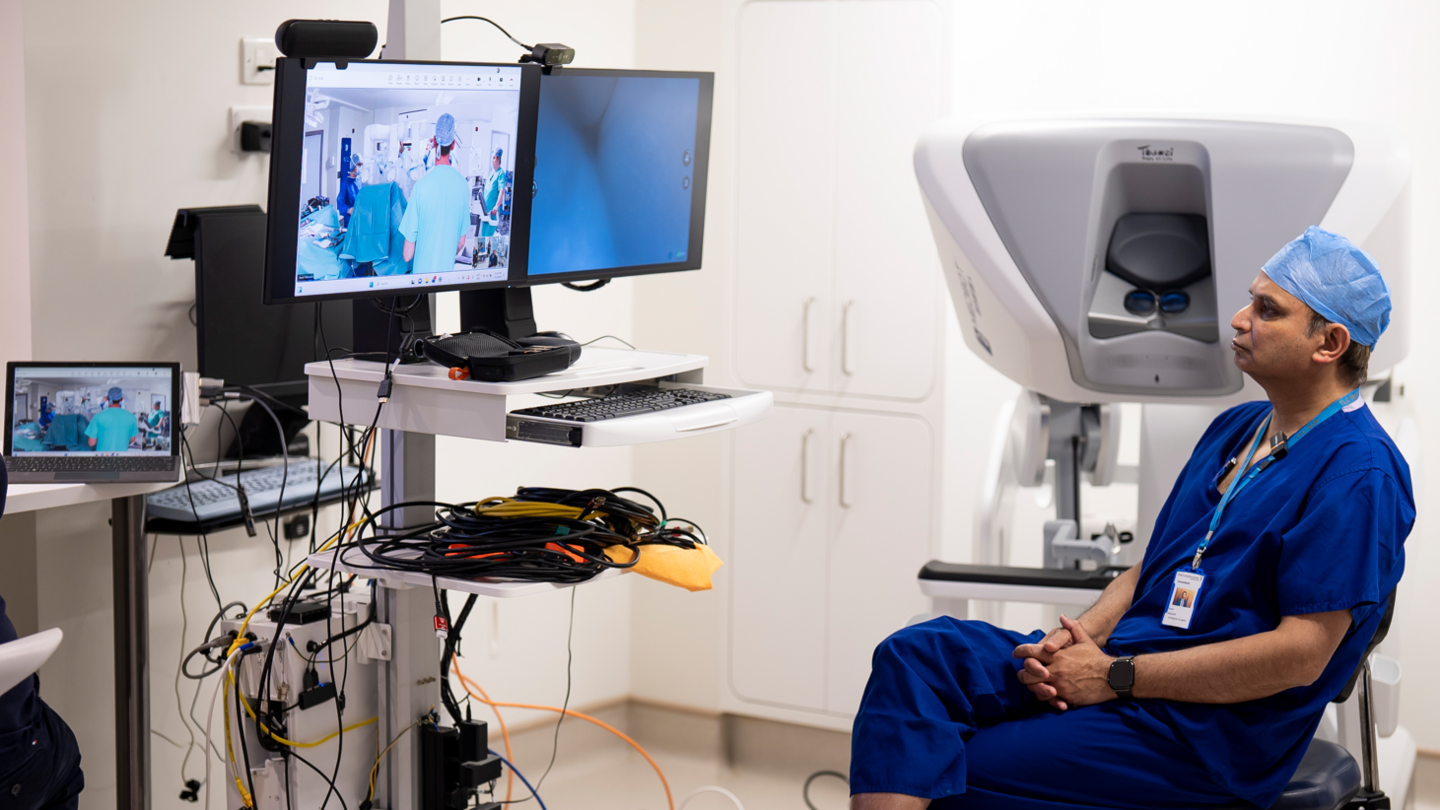 A doctor in blue sits looking at the two screens which show a surgery in another location. Lots of wires surround the display and a white box used for doing the surgery remotely can be seen behind him.
