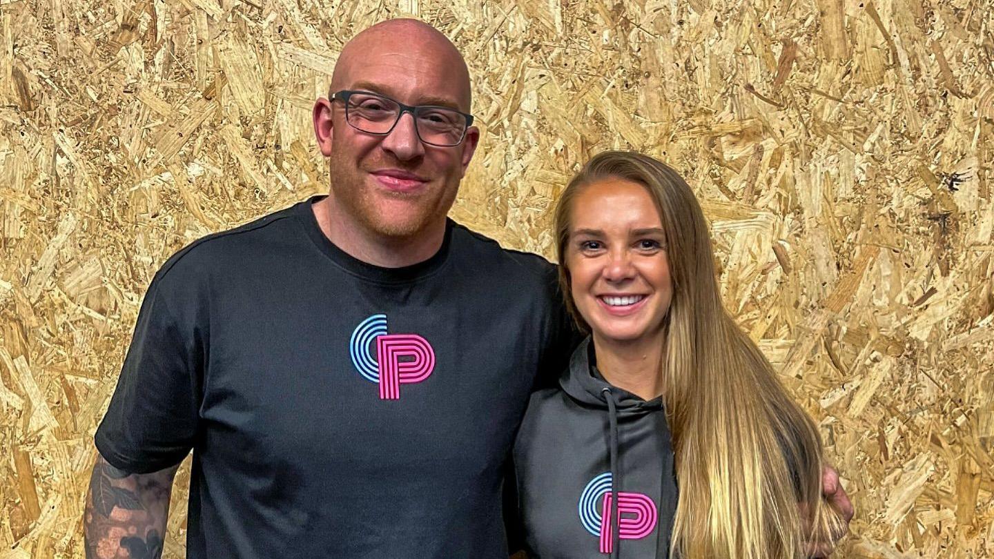 Mr Smart and Ms Blandford standing side by side, posing for the camera in front of a wooden chipping board in the background. They are wearing matching black merch from the gym, with a blue C and a pink P. Mr Smart is bald with a closely shaven beard, wearing dark framed rectangular glasses with a tattoo sleeve on his right arm. His left arm is around Ms Blandford, who has straight blonde hair.