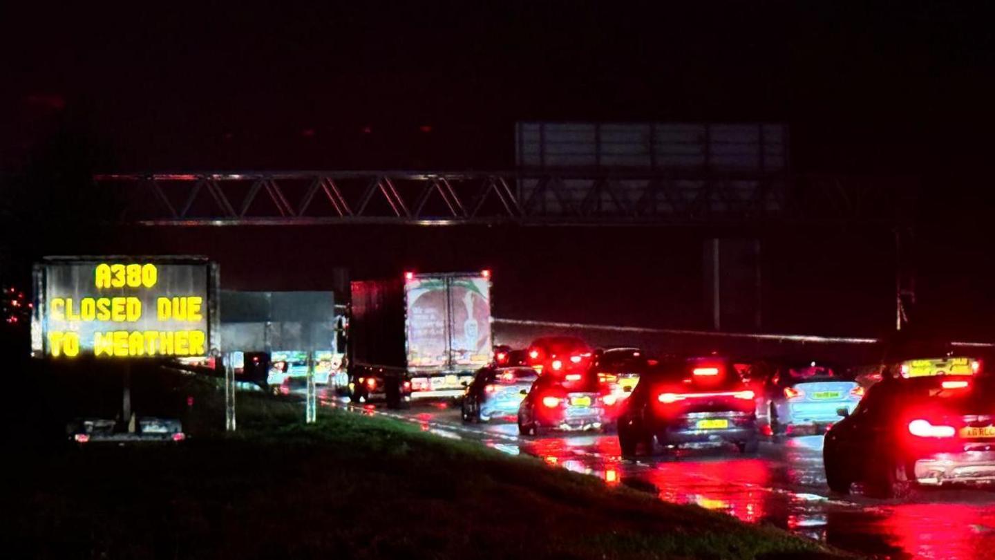 Standstill traffic on the A380. It is raining and car brake lights are glowing in the dark. A sign to the left of the road says 'A380 closed due to weather'.