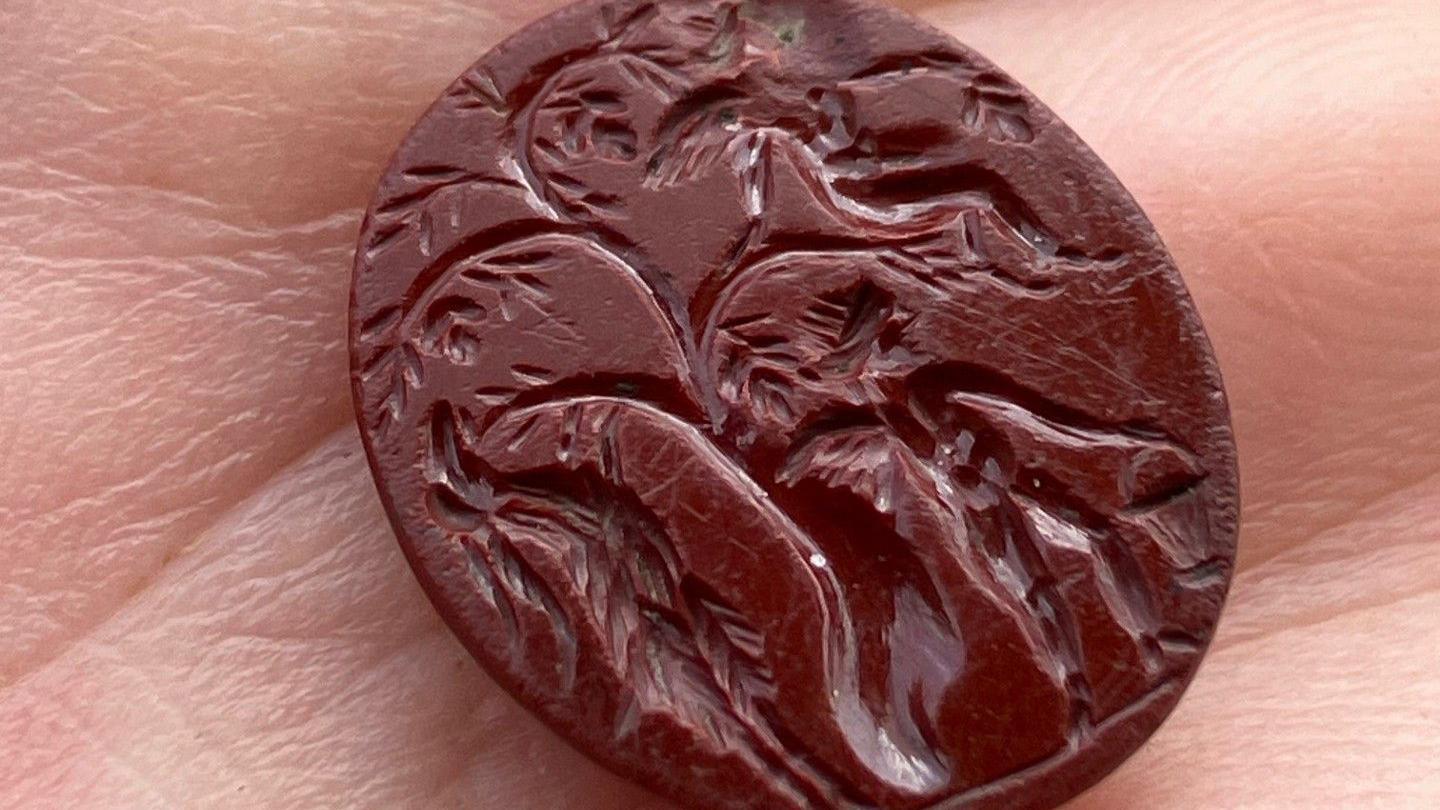 A red intaglio, probably a signet ring insert, which depicts two cupids picking grapes from a tree and a goat-like creature beside them.