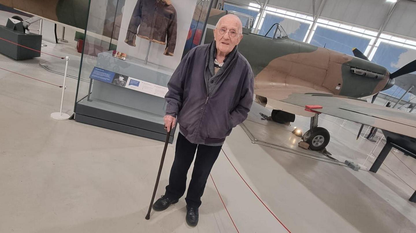 John standing in front of the aircraft display. He is wearing a grey jacket and holding his walking stick. The Spitfire is in the background, along with a display exhibiting a flying jacket.