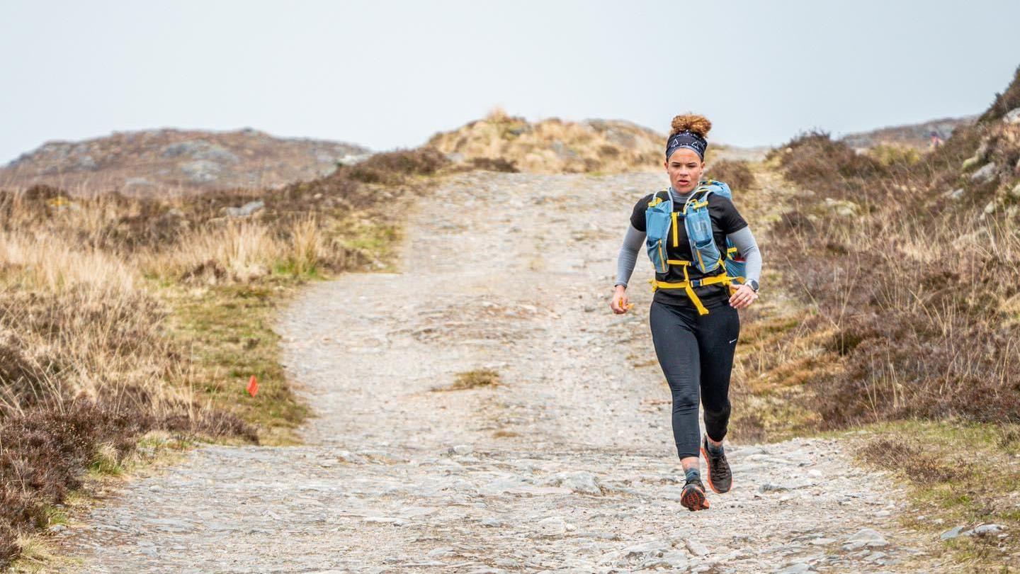 Sabrina Pace-Humphreys running on a trail in black leggings and a blue support bodypack. Her hair is in a high ponytail with a bandana. It is dry and moor-like behind her.