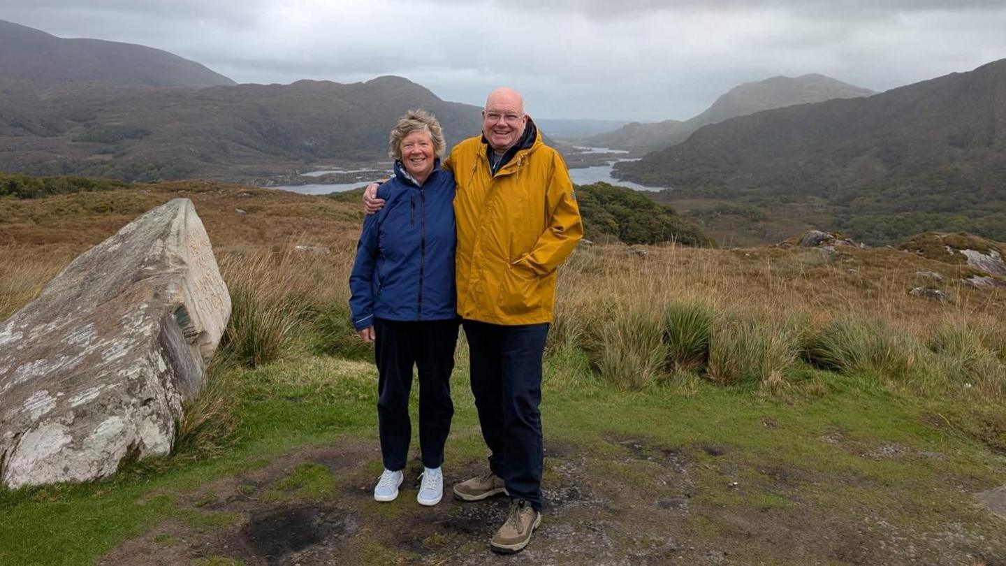 Older woman with grey hair in blue jacket and an older man in a yellow jacket smiling together with a rural backdrop including a lake and hills.