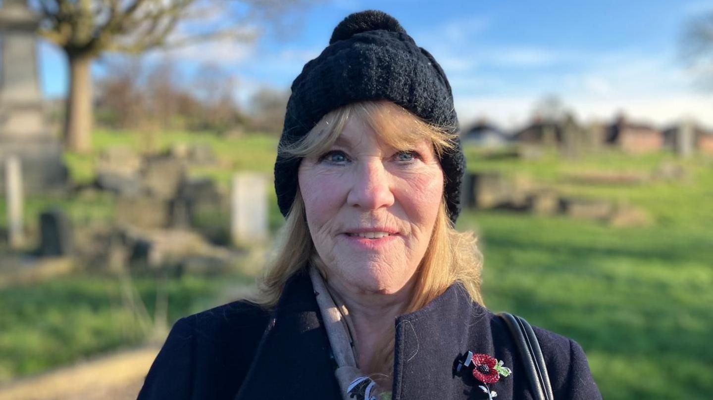 Christine Leeman looks at the camera, she is wearing a black wool hat and a poppy in her black blazer.