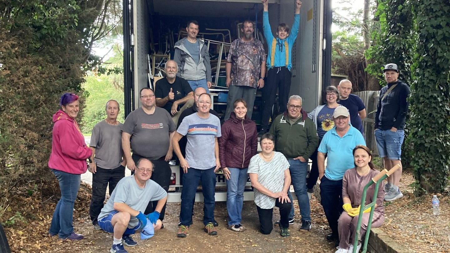 A group of volunteers standing in and in front of the back of a lorry. Furniture is visible in the lorry. There are hedges either side.