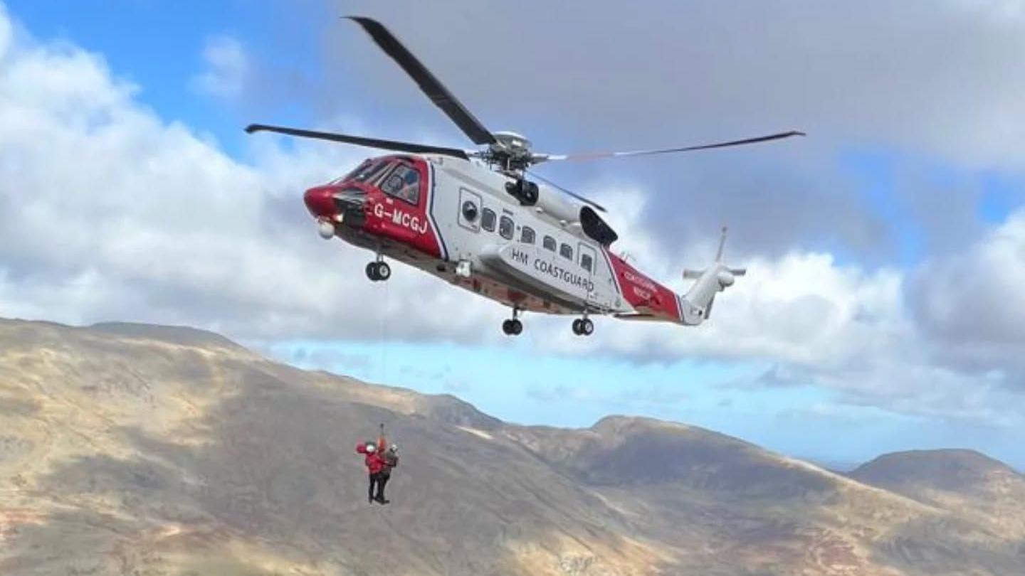 A red and white coastguard helicopter lifting two people in the air on a winch from the mountains in Eryri National Park. The sky is blue with patches of sun hitting the grassy mountain range in the background. 