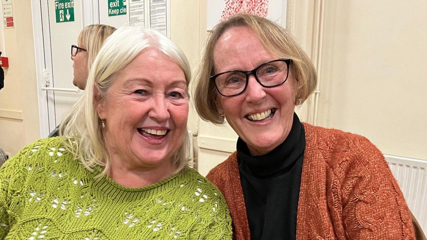 Two middle-aged women in jumpers, leaning their heads towards each other for the photo, both smiling broadly.