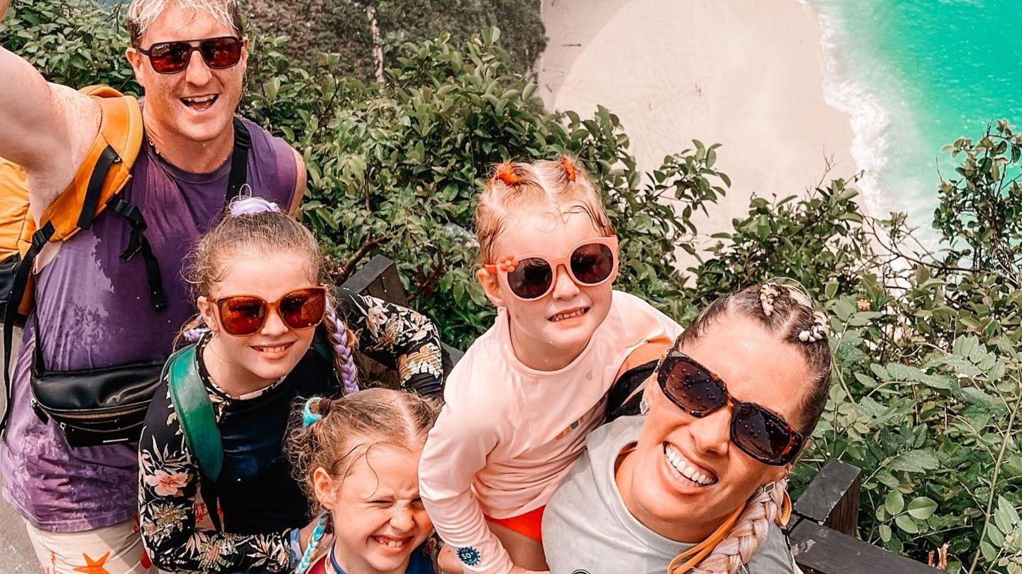 The family smile at the camera at a clifftop view point, with the beach and turquoise sea visible below